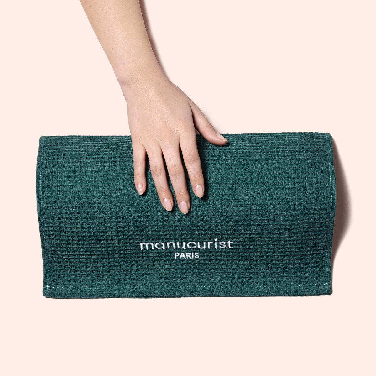 A manicured hand rests on a folded, textured green Manucurist-us Towel, embroidered with the brand name, providing gentle surface protection against a pale pink background.