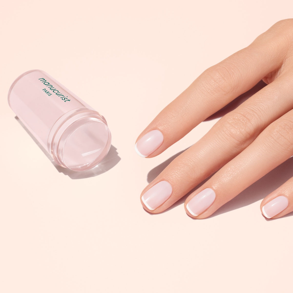 A hand with neatly manicured, flawless French tips rests next to a translucent pink Mani stamper by Manucurist US on a light pink surface.