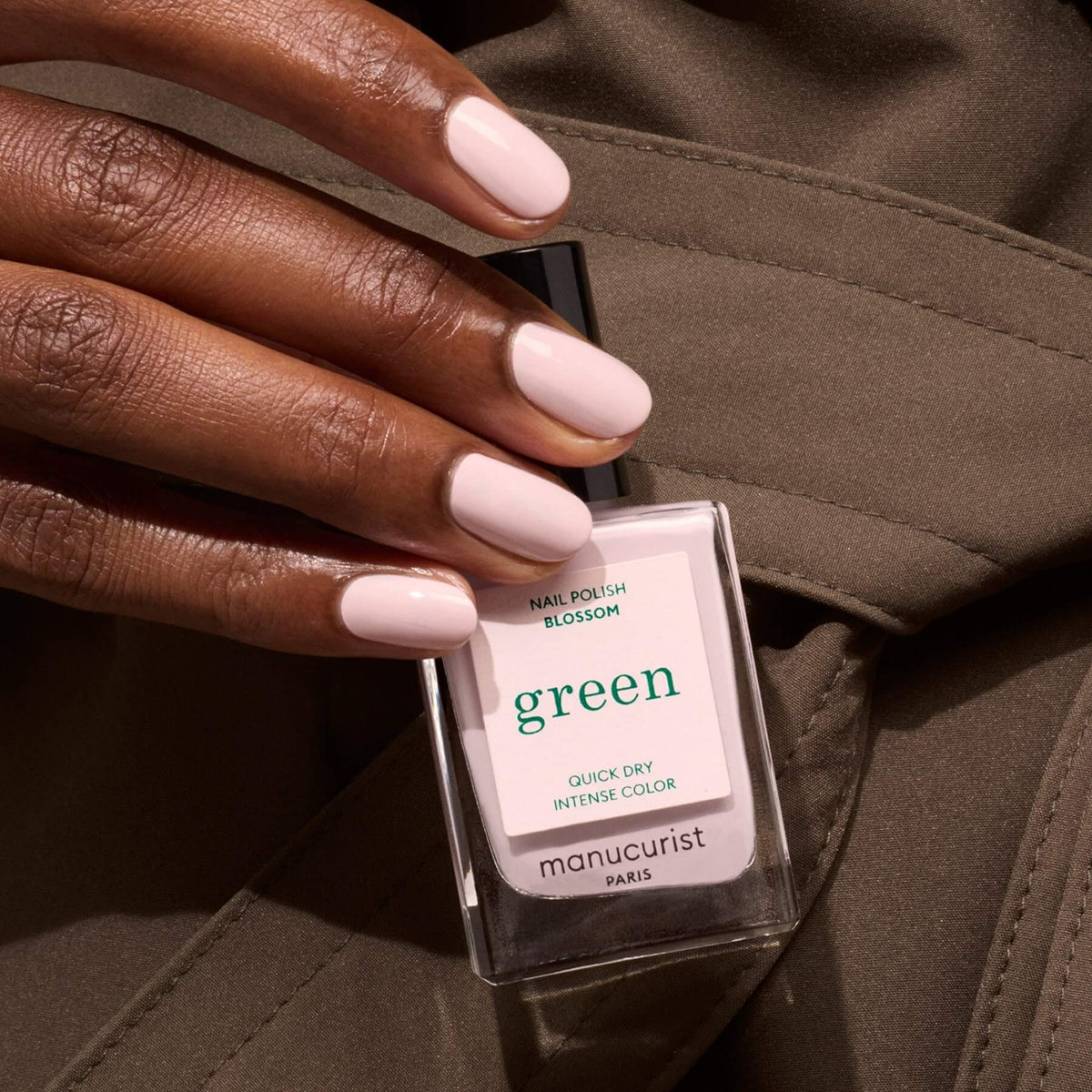 A hand with neatly manicured, pale pink nails holds a bottle of Manucurist Blossom nail polish, set against olive-green fabric.