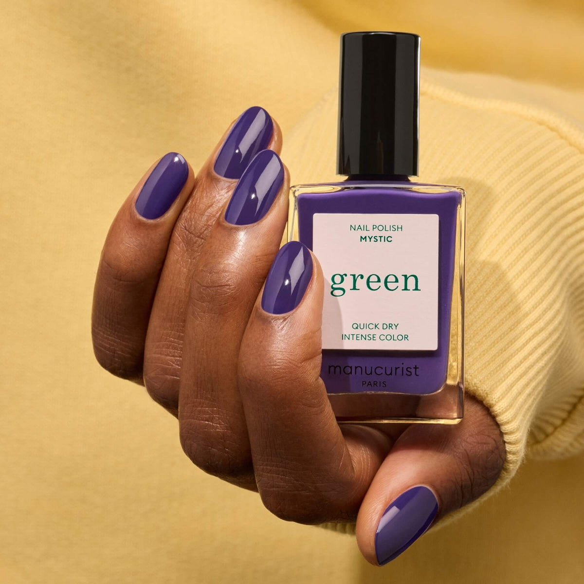 A hand with neatly manicured dark violet nail polish holds a bottle labeled Mystic by Manucurist against a yellow background.