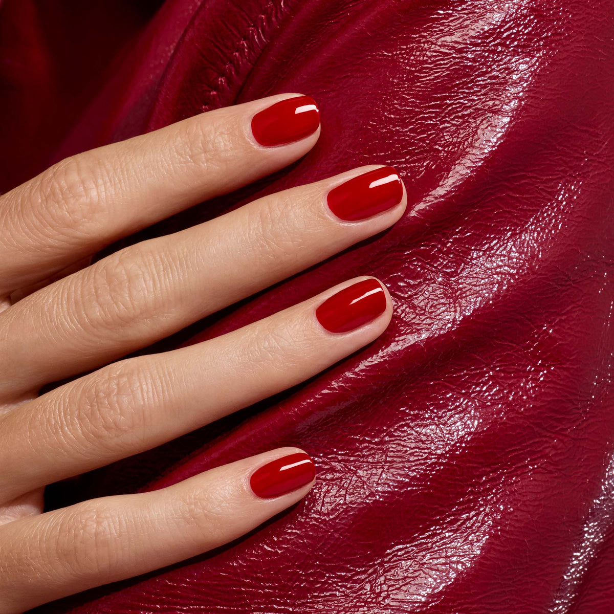 Gros plan sur une main aux ongles impeccablement manucurés avec le vernis Red Velvet de Manucurist, posée sur une surface rouge brillante et texturée.