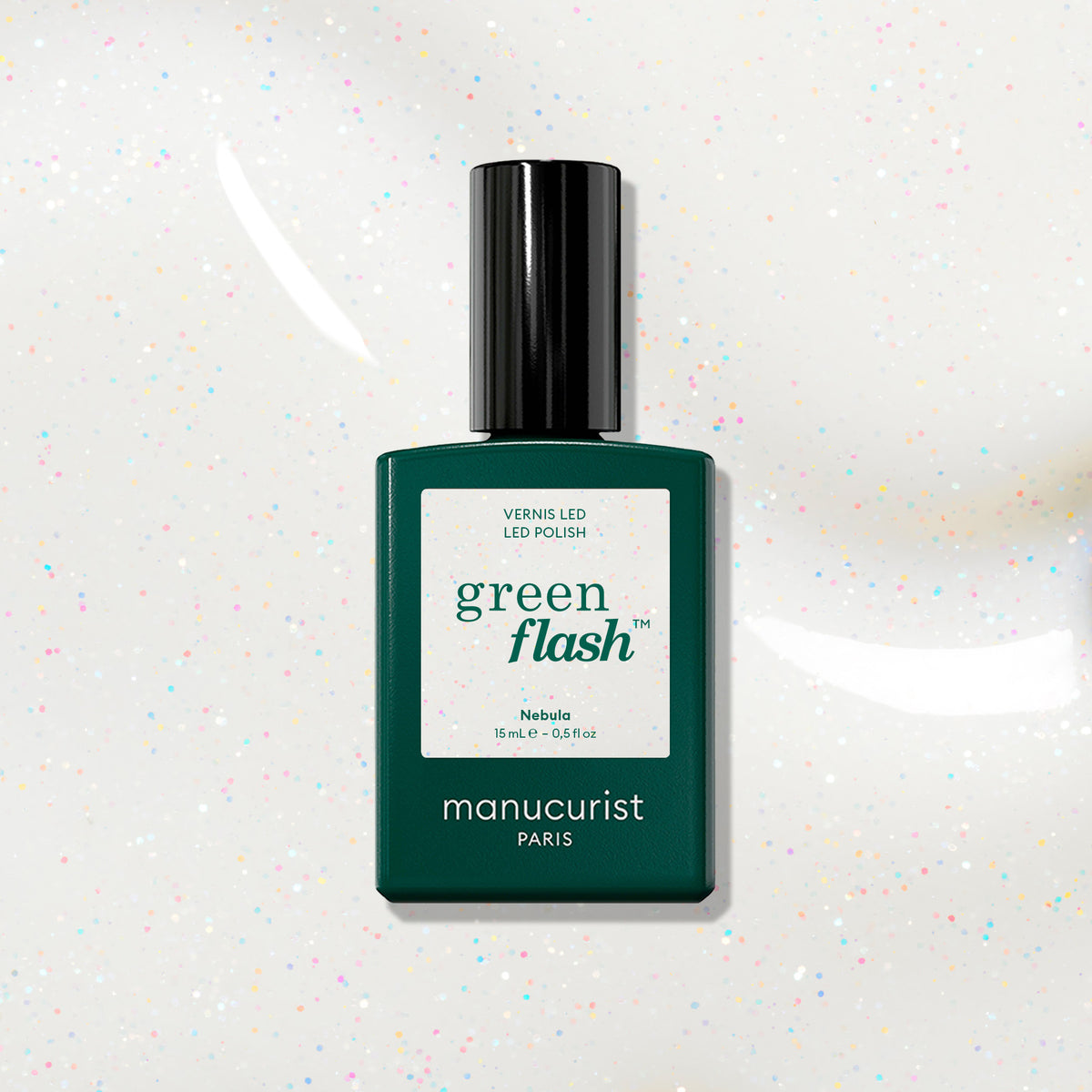 A bottle of Manucurist Nebula nail polish rests on a white surface with colorful specks. The green bottle, labeled Nebula, suggests a holographic effect under its sleek black cap.