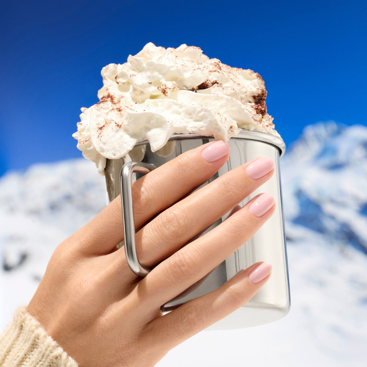 A hand with Manucurist’s Milky Pink nail polish holds a metal mug of hot chocolate topped with whipped cream, set against snowy mountains and a bright blue sky.