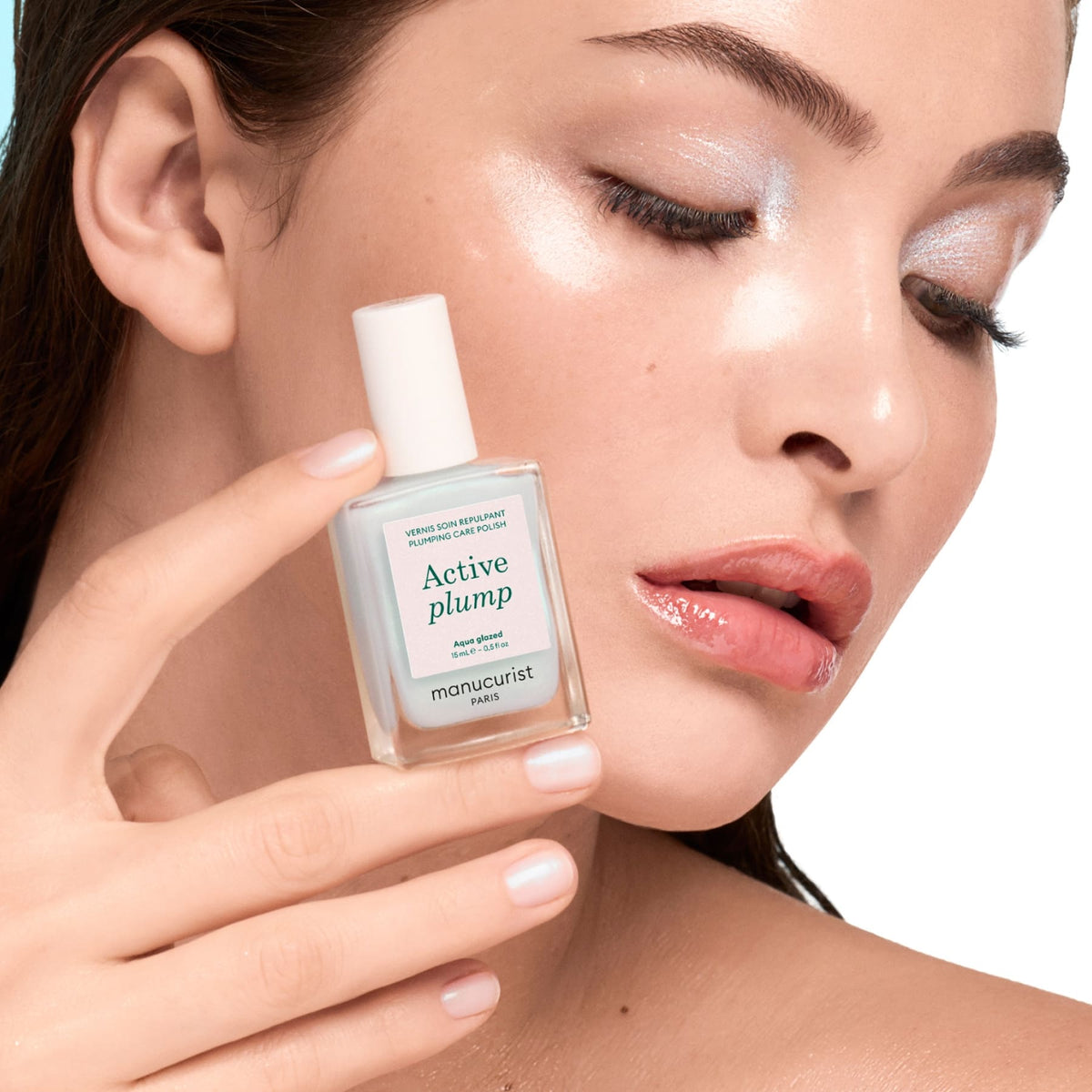 A woman with dewy makeup holds a bottle labeled Active™ Plump Aqua Glazed by Manucurist close to her face, highlighting her glowing, radiant skin and shimmery eyelids. Known for its volumizing nail care benefits, this product also promotes beautifully hydrated nails.
