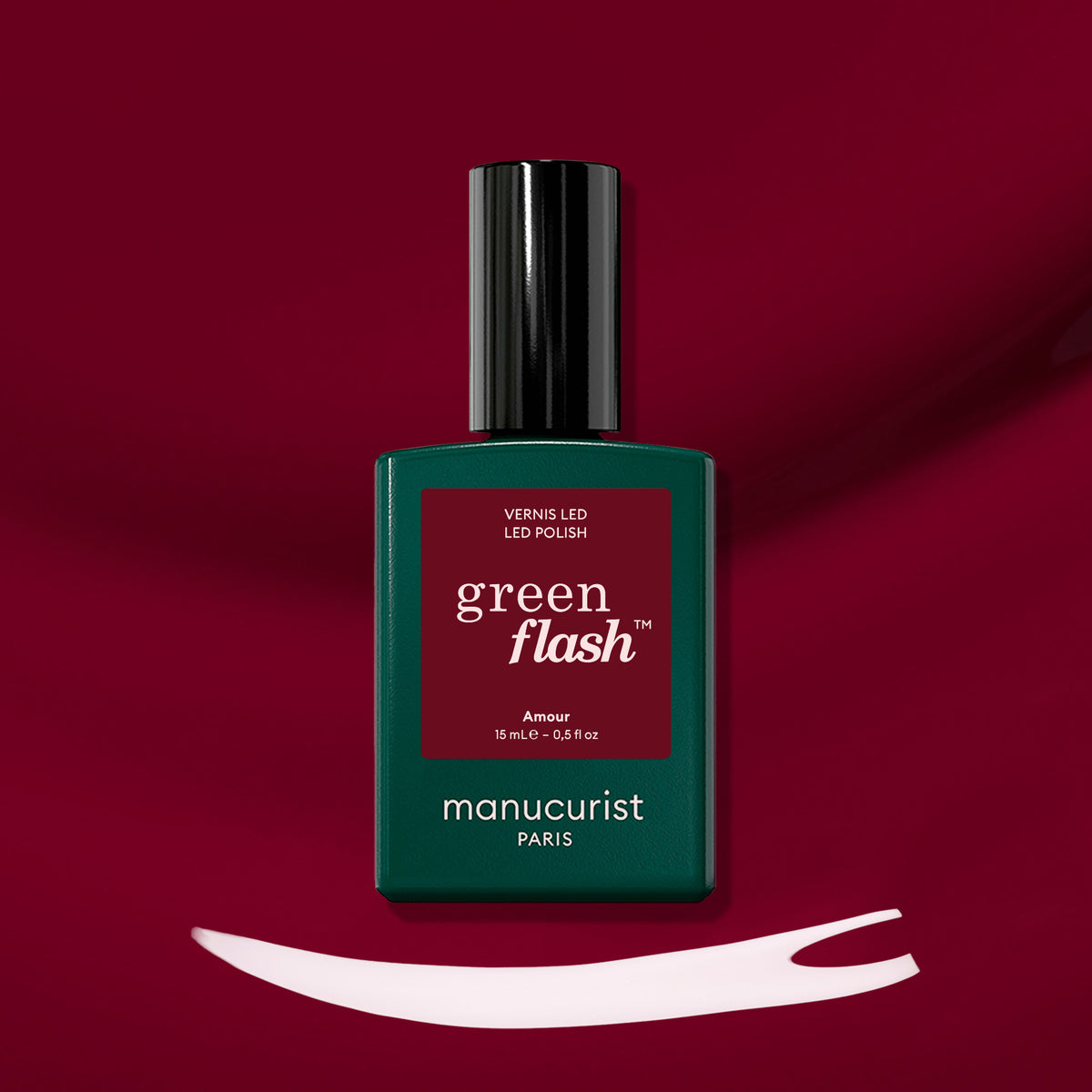 A bottle of Amour LED nail polish by Manucurist rests on a deep red surface with a brush stroke of white polish below it, highlighting the luminous burgundy hue of this signature gel polish.