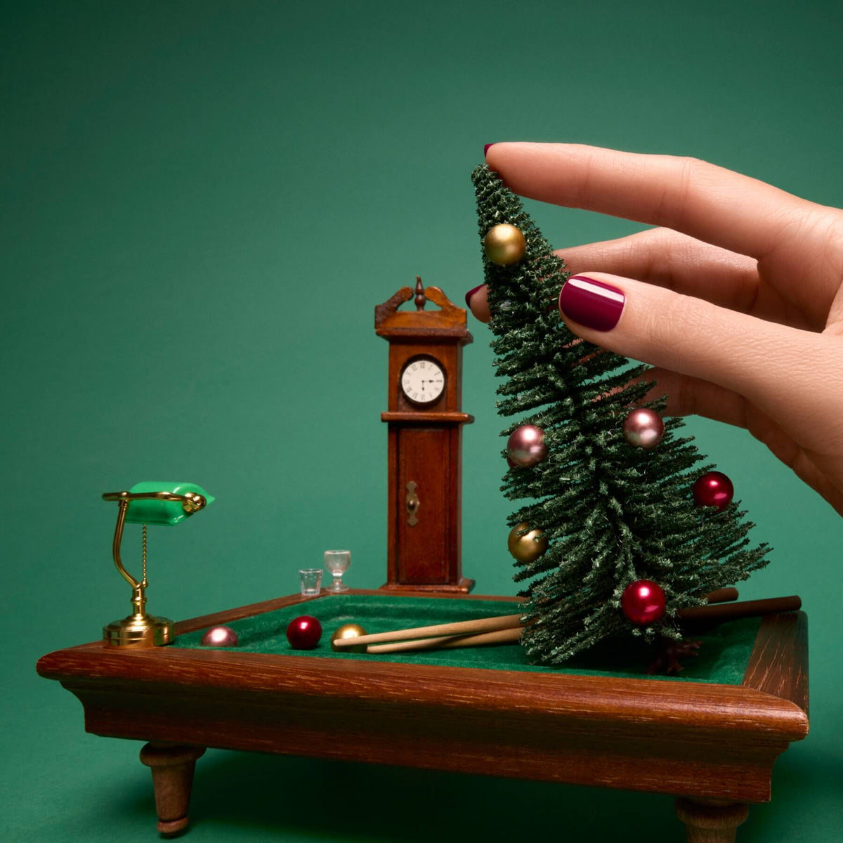 A hand adorned with Manucurist’s Amour luminous burgundy nail polish places a miniature Christmas tree on a small green pool table. In the background, a tiny lamp, two cue sticks, and a clock are visible against a green backdrop inspired by the Green™ range.