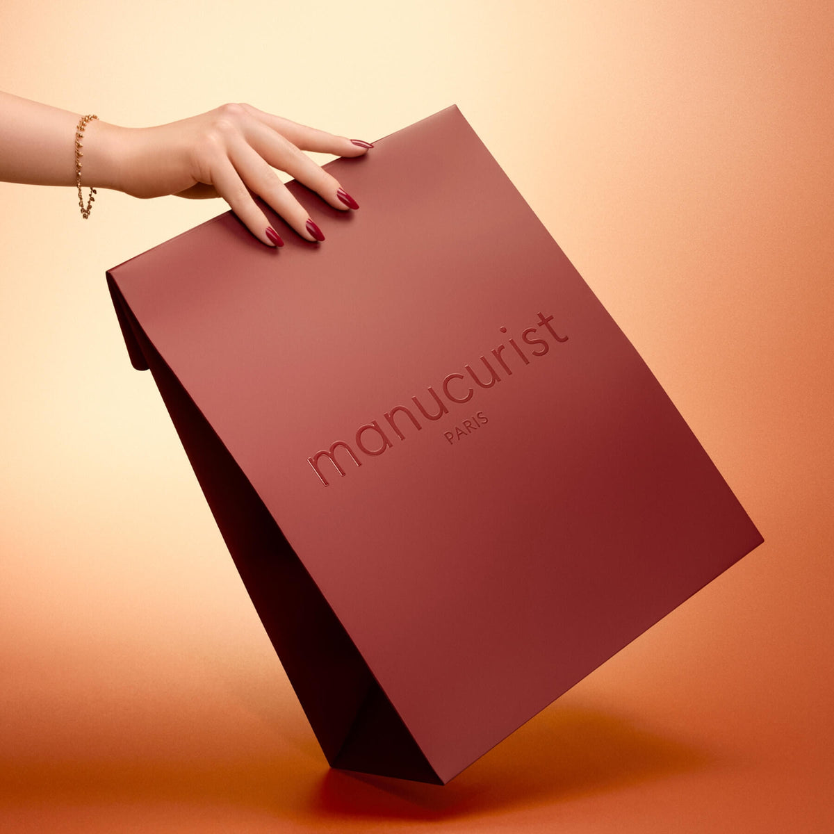 A hand with painted nails and a bracelet holds the large Holiday Gift Bag - M from Manucurist, featuring a matte burgundy finish, against a gradient peach background.