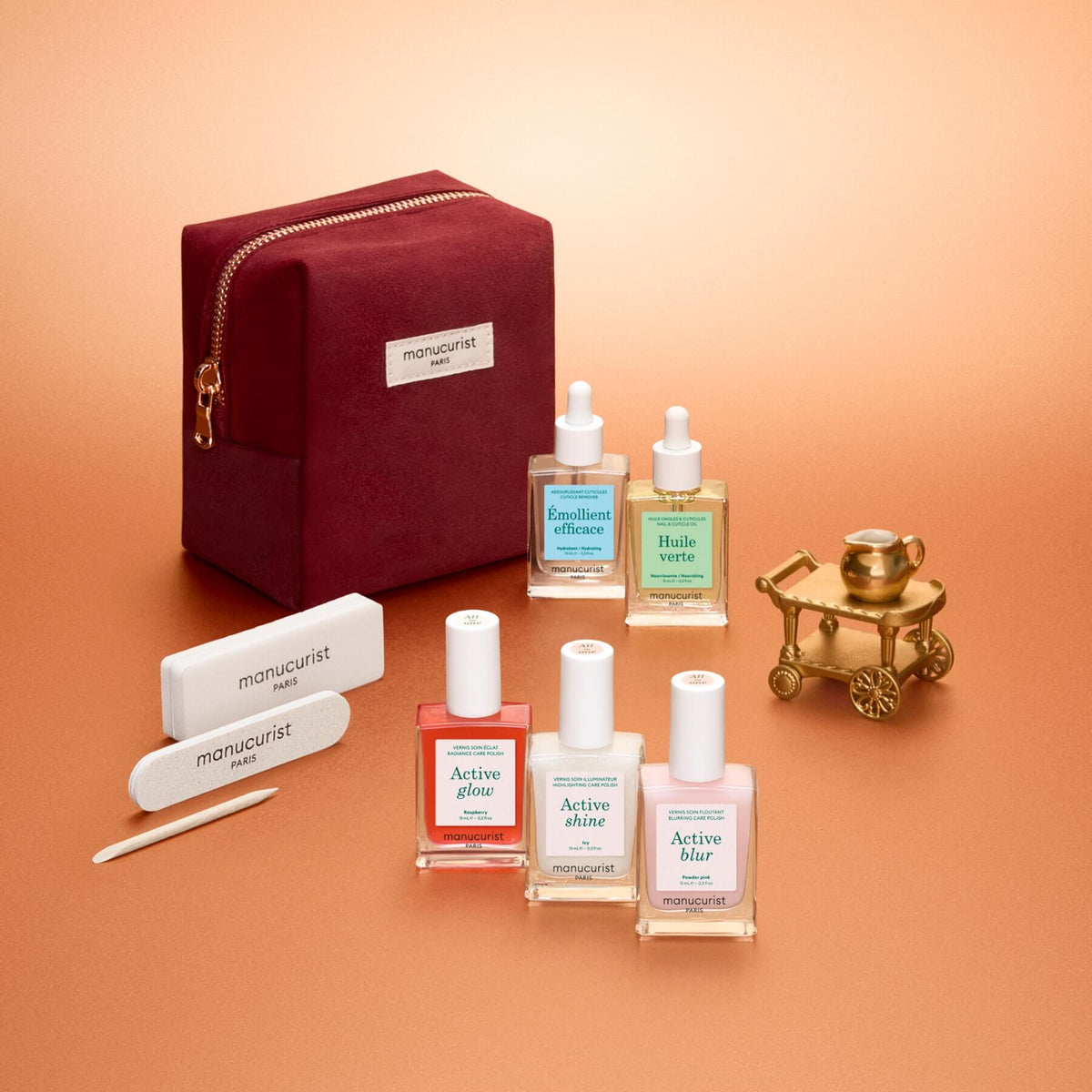 The Manucurist Active™ Starter Kit Gift Set, featuring nail care bottles, nail files, a cuticle stick, and a red zippered pouch elegantly presented with a decorative gold cart on a peach-colored background, is the perfect holiday gift for anyone who loves maintaining beautiful nails as part of their manicure routine.