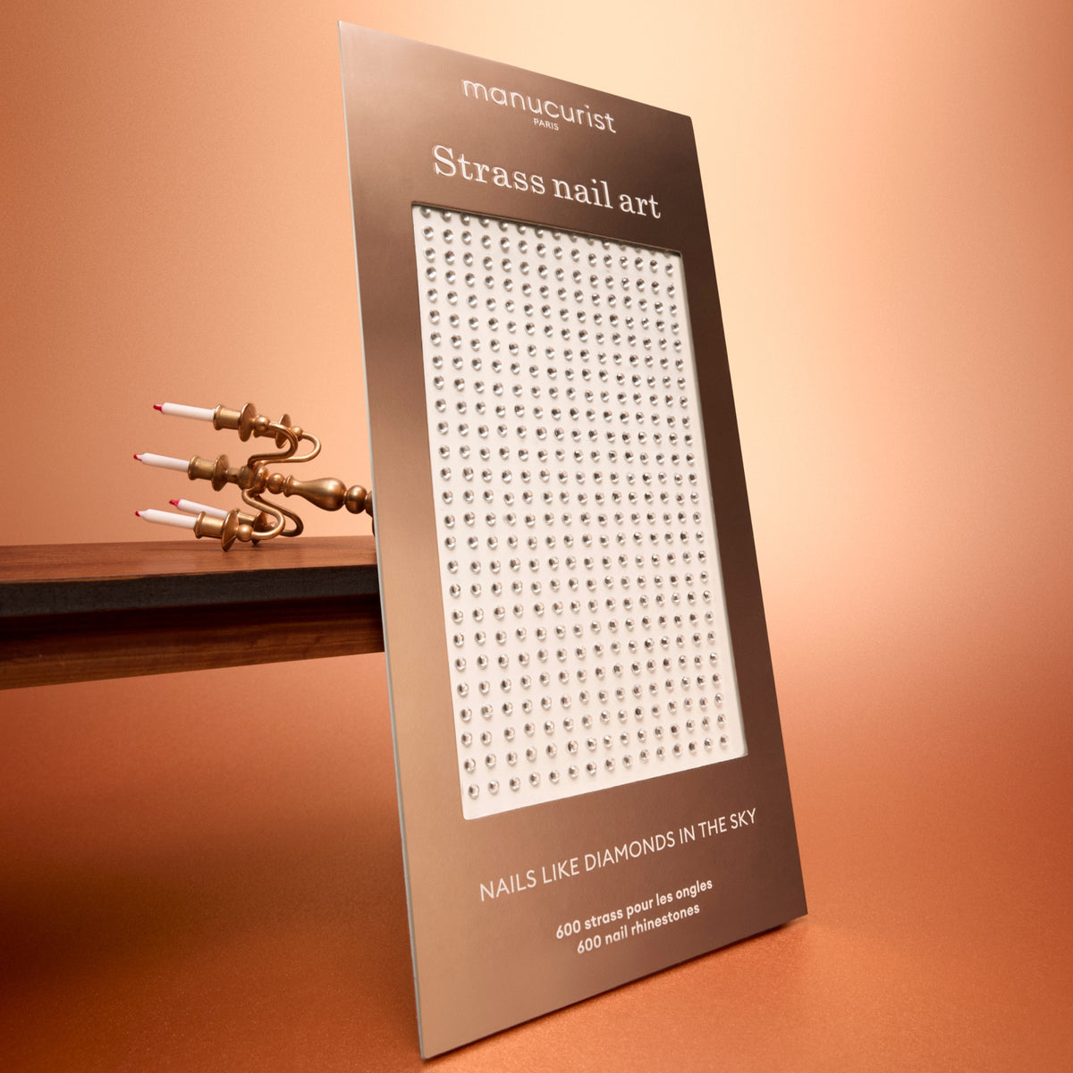 Un paquet de strass Nail Art Holidays de Manucurist est présenté debout sur une surface marron, avec un bougeoir doré moderne en arrière-plan. Idéal pour sublimer votre prochaine manucure ou lui apporter une touche festive, il présente des strass argentés alignés en rangées régulières, comme sur une planche de strass pour nail art professionnel.