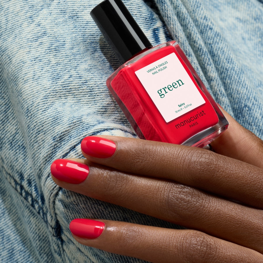 A hand with neatly manicured nails painted Spicy, a red shade by Manucurist, holds a matching bottle of Manucurist nail polish from the Green™ range against light blue denim fabric.