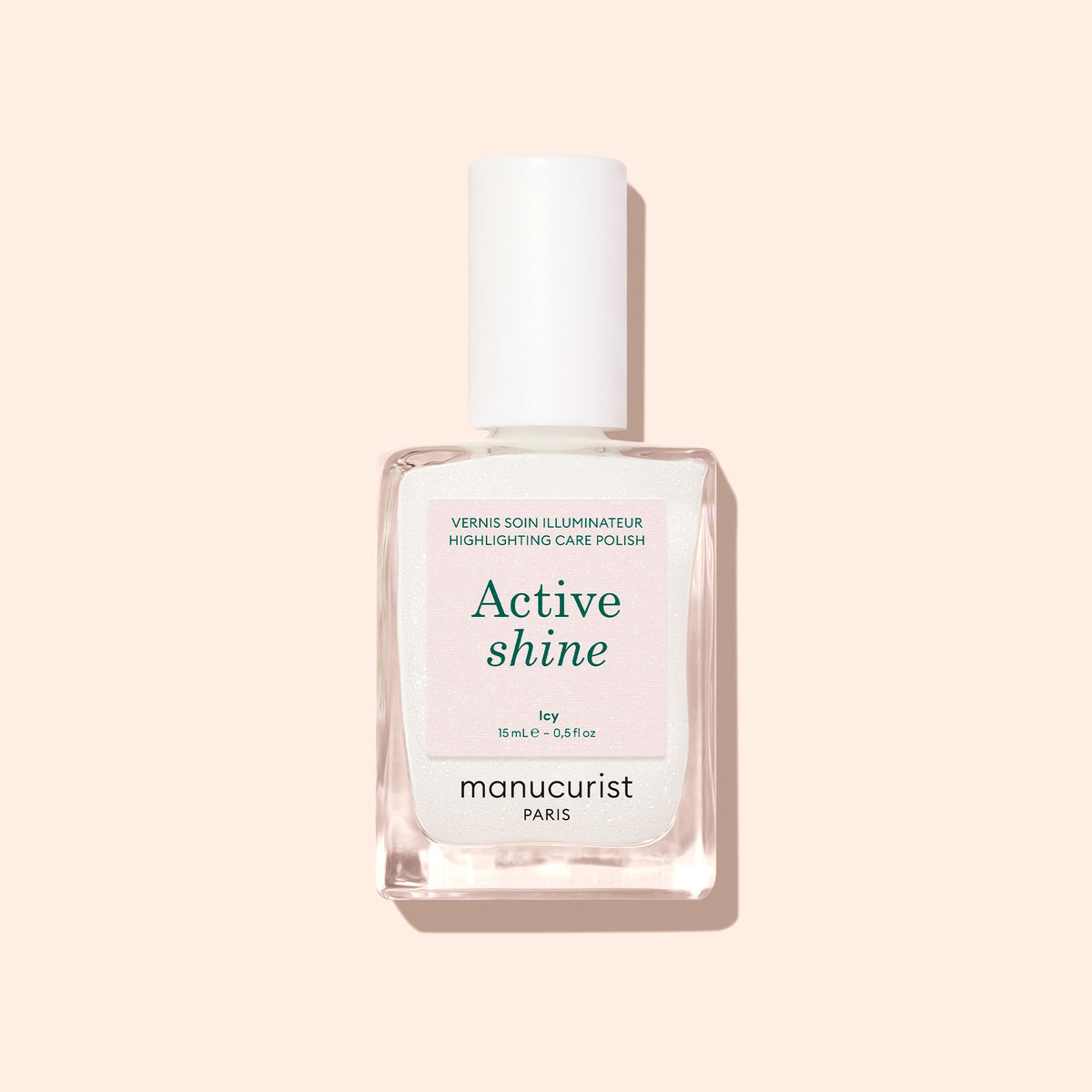A clear glass bottle of Manucurist Active™ Shine Icy illuminating nail polish with a white cap, photographed against a pale peach background.