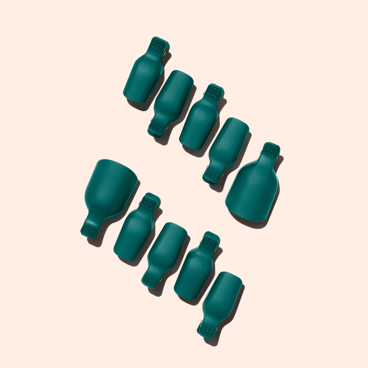 Nine teal Foot Removal Clips by Manucurist, shaped like bottles, are arranged diagonally on a light beige background. Each casts a shadow to the left, with two clips noticeably larger than the others.