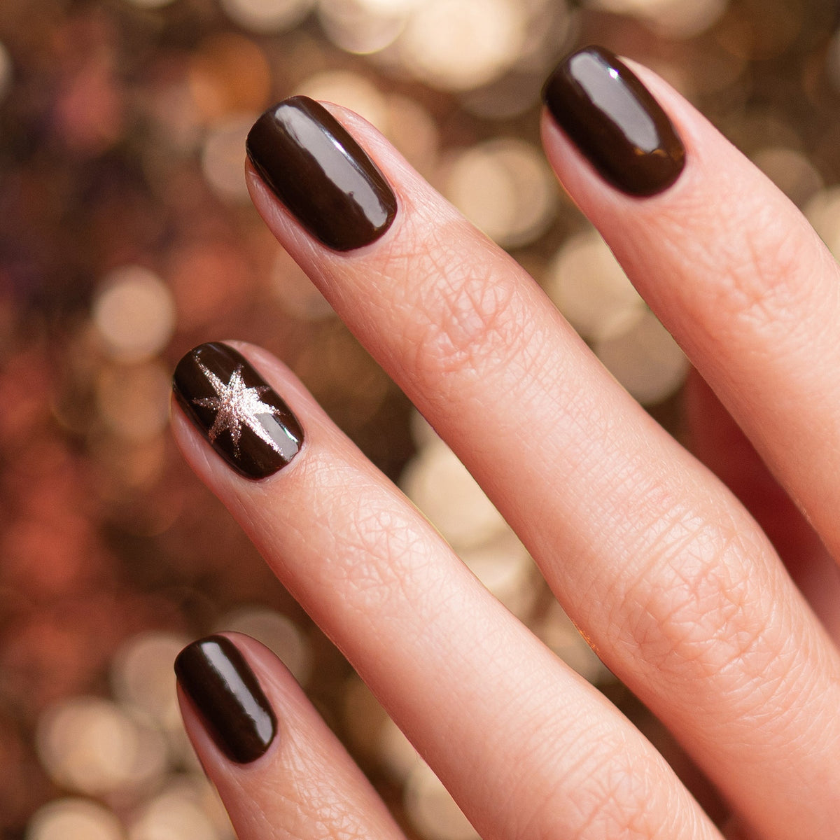 A hand showcases Manucurist’s Clove, a glossy chocolate brown polish, with a small silver star on the ring fingernail. The Green™ range color stands out against a blurred background of warm golden lights.