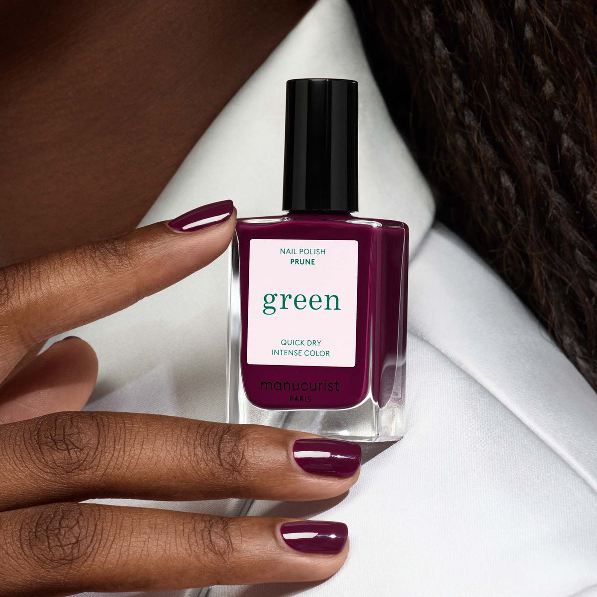 Une main à la peau foncée présente des ongles soigneusement manucurés avec le vernis Manucurist Prune, un vernis violet foncé à séchage rapide, tout en tenant le flacon assorti sur fond blanc.