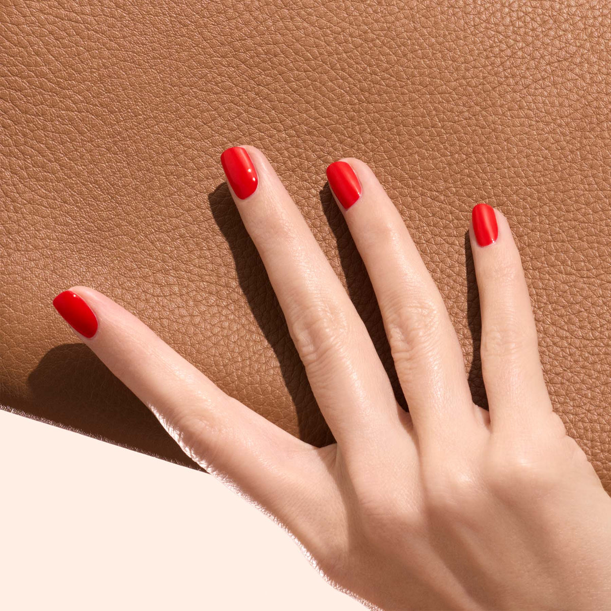 A hand with neatly manicured nails painted in Manucurist’s Anémone, a vibrant red shade, rests on a textured brown surface against a soft, neutral background.