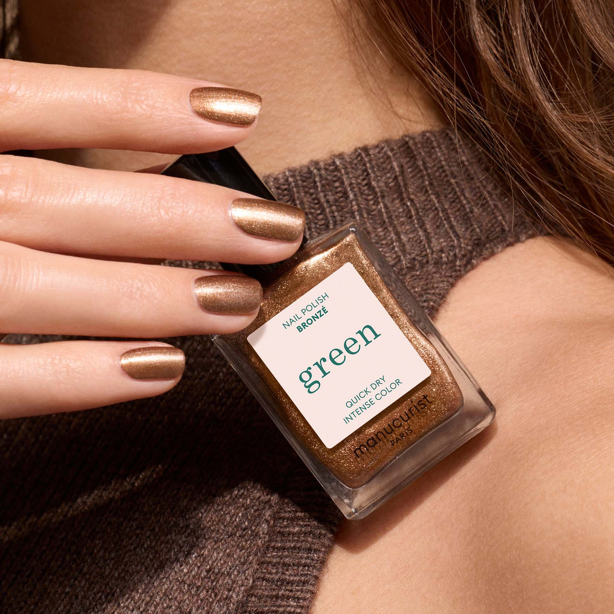 A hand with Bronzé, a bronze metallic nail polish by Manucurist, holds a matching bottle labeled "Manucurist Bronzé" against a brown knit sweater background.