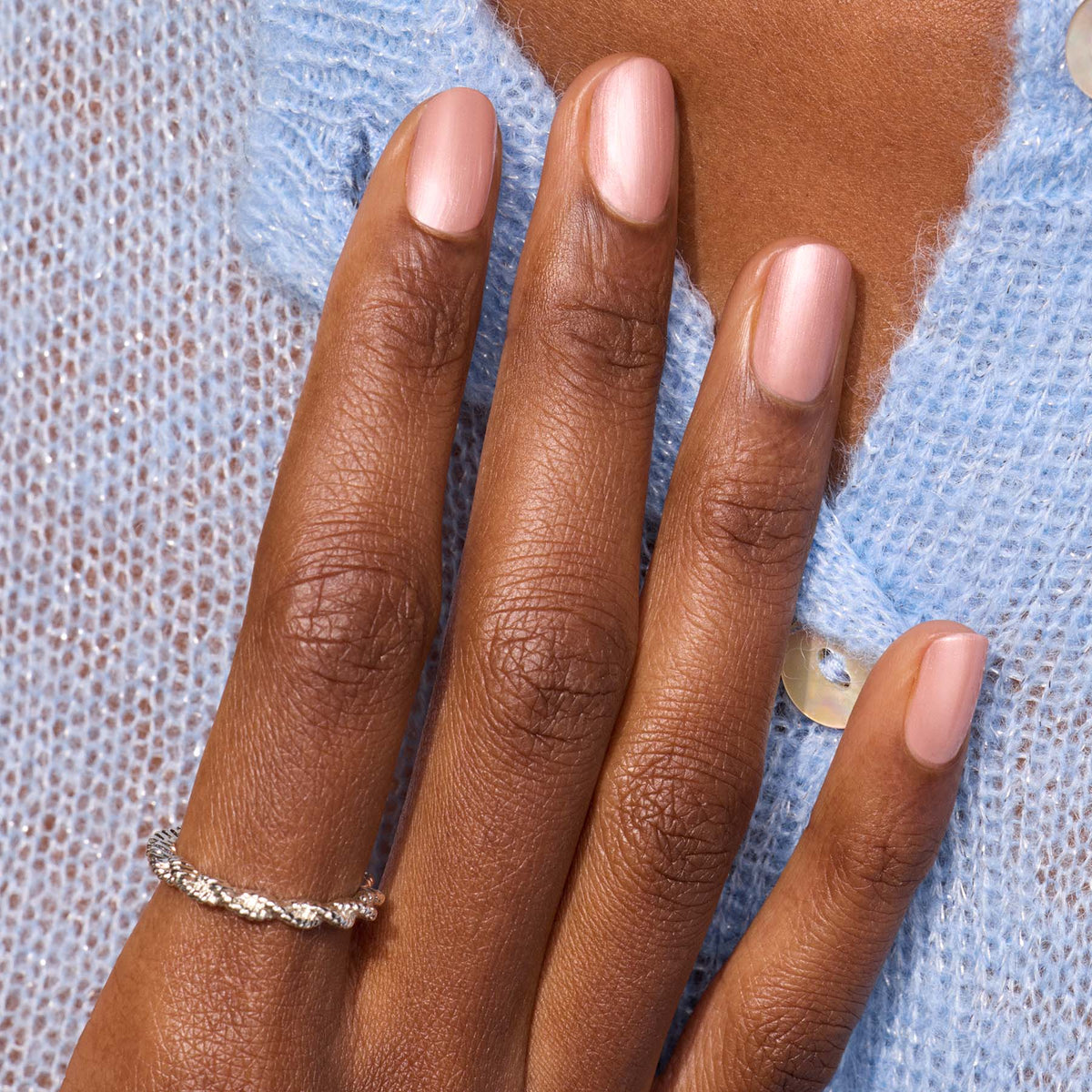 A hand with neatly manicured Carnation gel polish by Manucurist wears a delicate twisted silver ring on the ring finger, resting on a light blue textured knit sweater with white buttons.