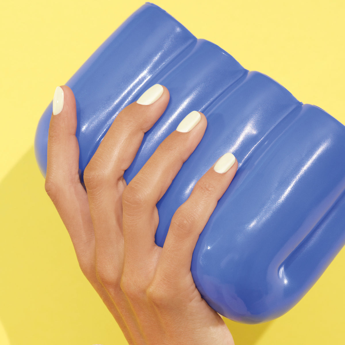 Une main à la manucure soignée, vernie de la teinte crémeuse Porcelaine de Manucurist, tient un objet arrondi bleu brillant sur un fond jaune vif.