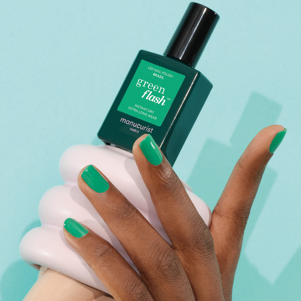 Une main aux ongles soigneusement manucurés et peints en vert électrique tient un flacon de vernis Brazil Manucurist sur un fond bleu clair.