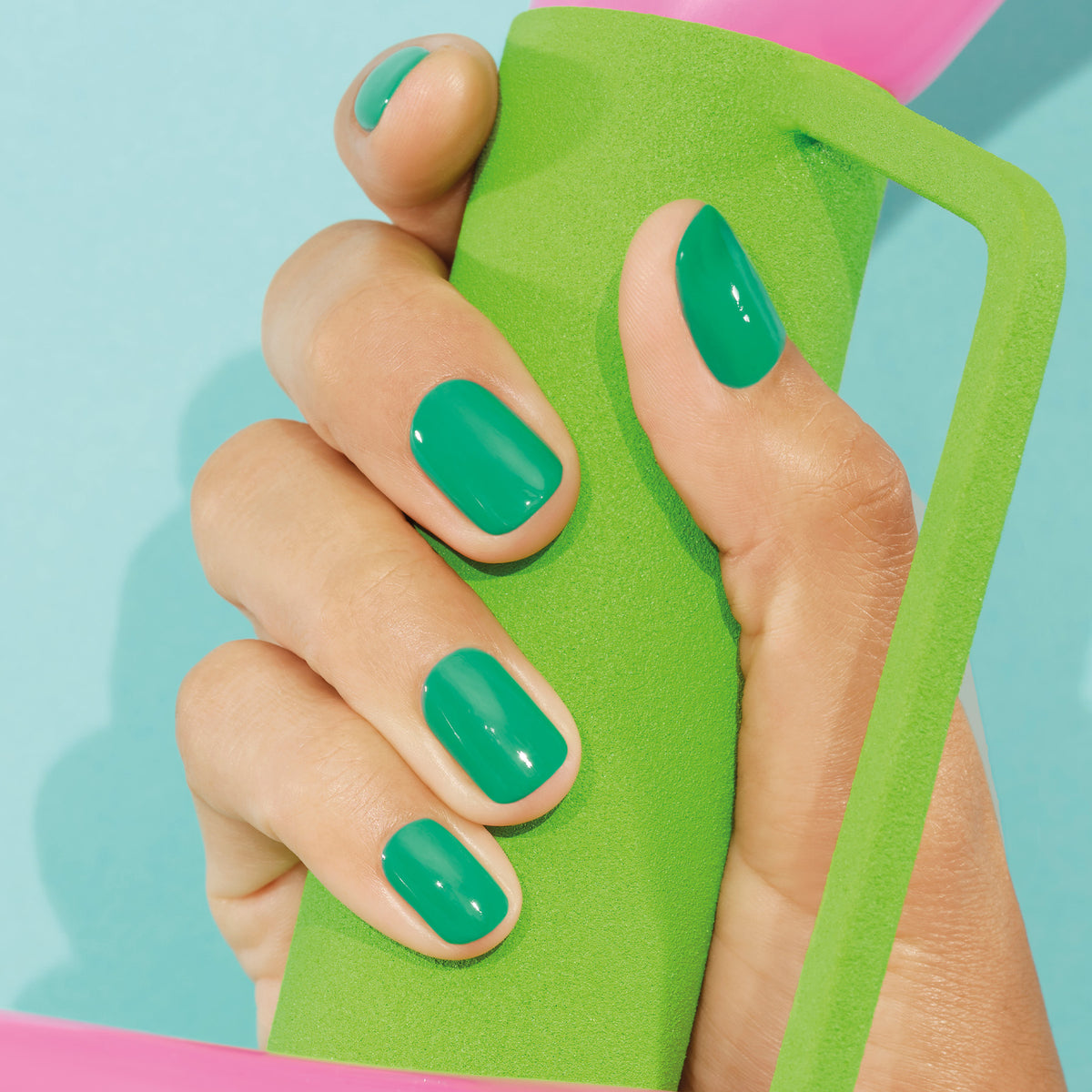 A hand with neatly manicured nails painted in Manucurist’s Brazil, an electric green shade, and finished with Top Coat holds a green foam handle against a light blue background.