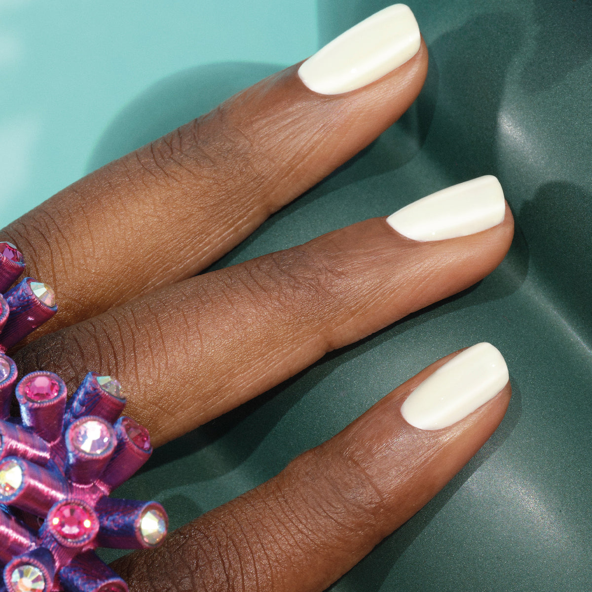 Gros plan sur une main à la peau foncée et lisse, aux ongles courts peints avec le vernis à ongles Porcelaine de Manucurist. La main porte une grosse bague violette ornée de gemmes rondes brillantes, sur un fond vert sarcelle.