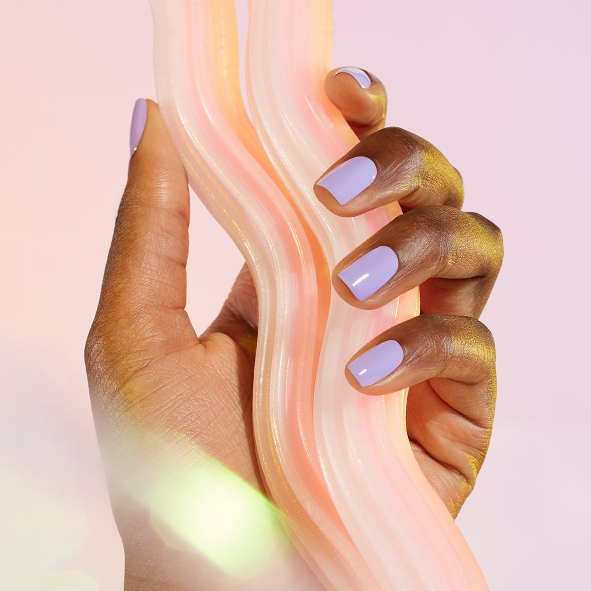 A hand with Manucurist’s Wisteria pastel purple nail polish holds a wavy, translucent object with cool blue undertones against a soft pink background.