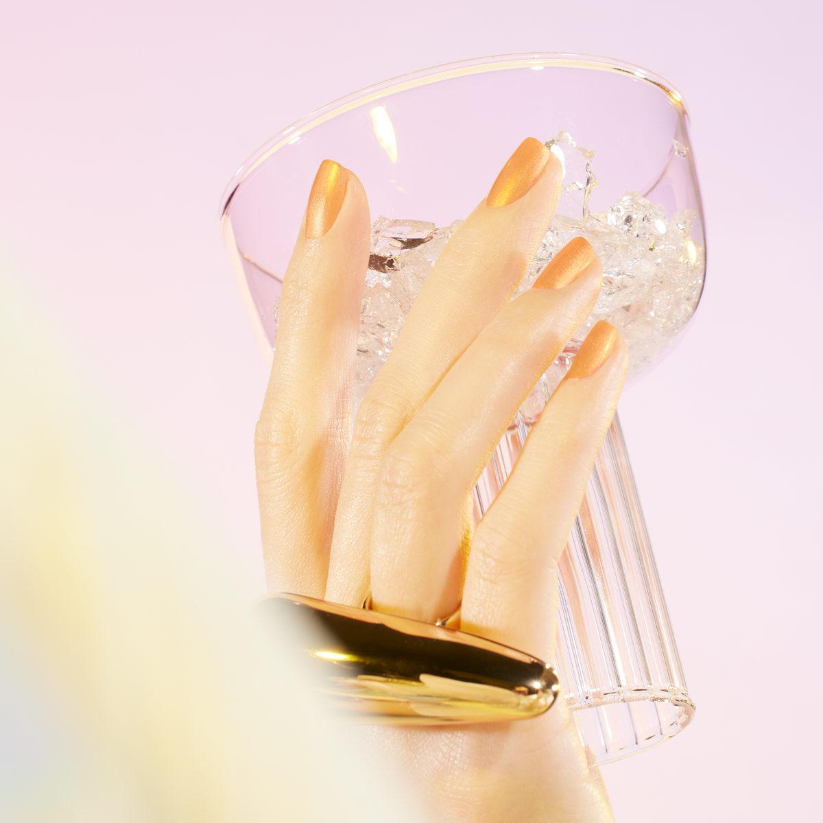 A hand with Manucurist’s Pamplemousse metallic gold nails and a chunky gold ring holds a clear glass of ice against a pastel background, the luxe look finished with pearlescent polish shimmer.