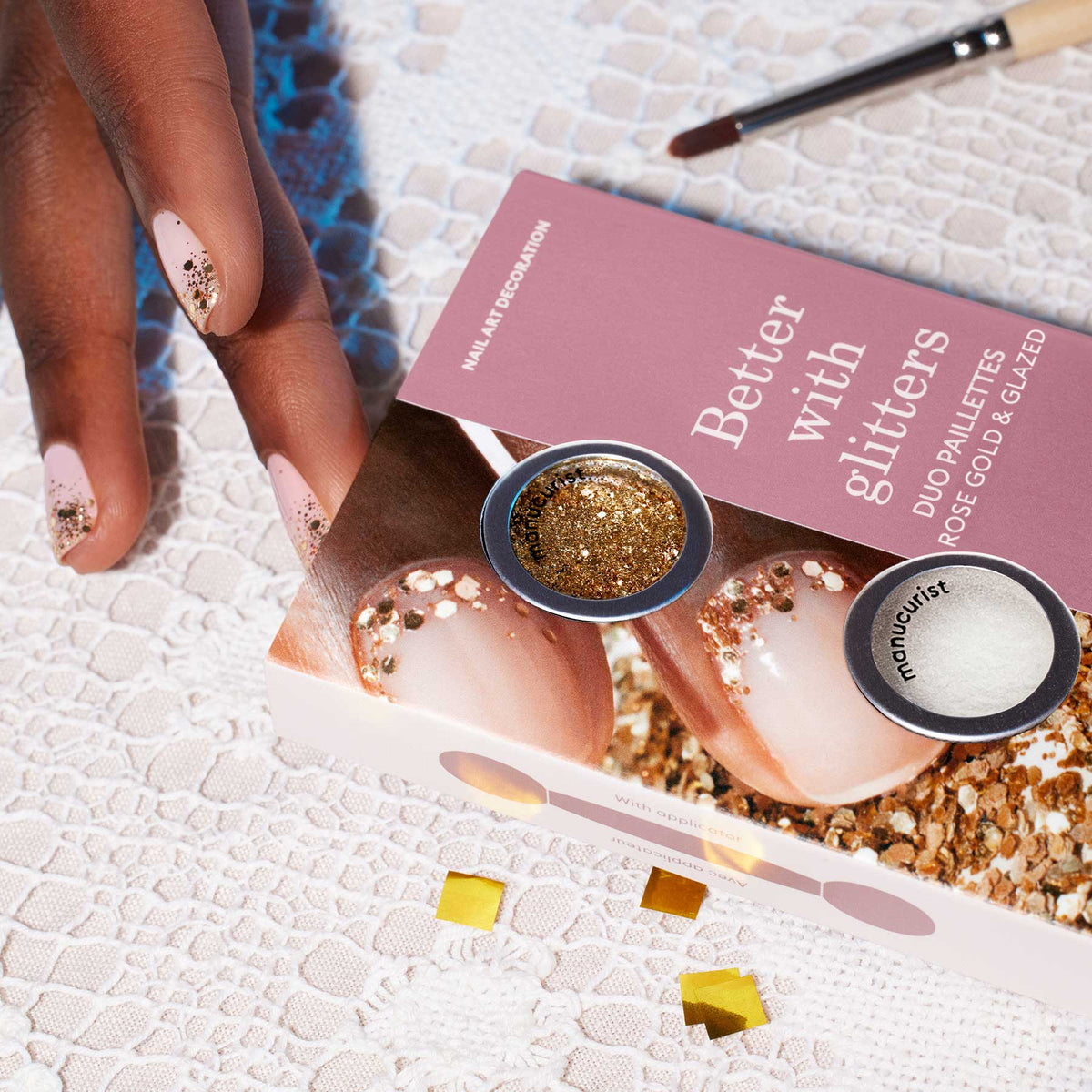 A hand with nude nails, adorned with Manucurist’s Glitters Duo Rose Gold Glazed, rests on lace beside a pink and white "Better with glitters" box, showcasing rose gold and pearl powders for a stunning pearlescent shine.