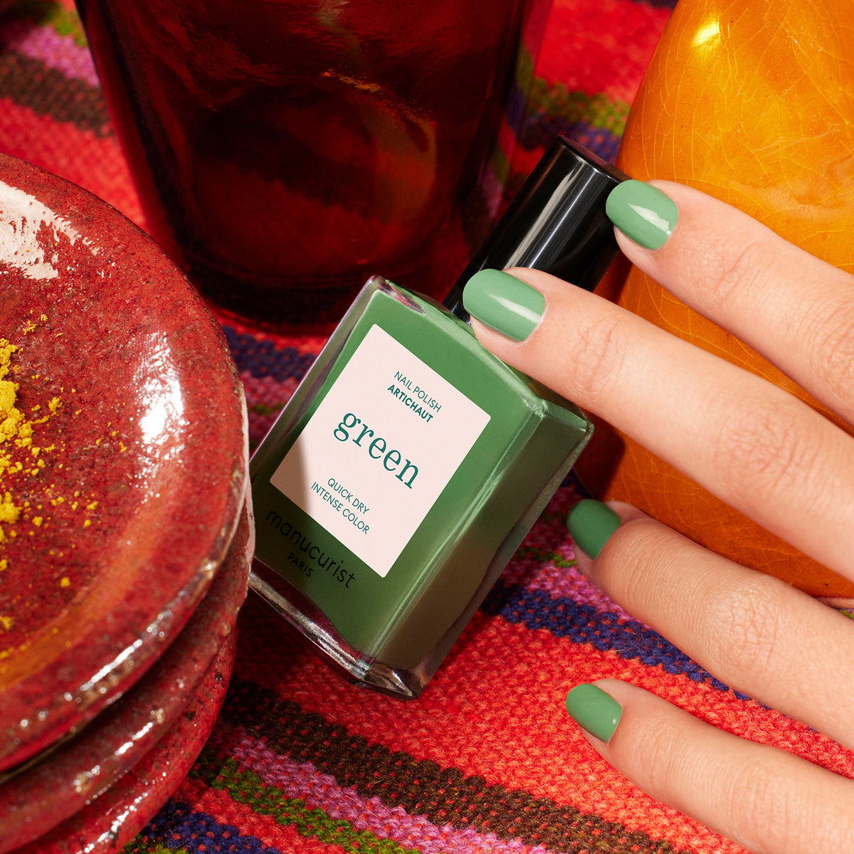 Une main aux ongles vernis de vert tient un flacon de vernis à ongles Manucurist étiqueté Artichoke, posé sur un tissu tissé vibrant et entouré de céramiques rouges et orange rappelant l&