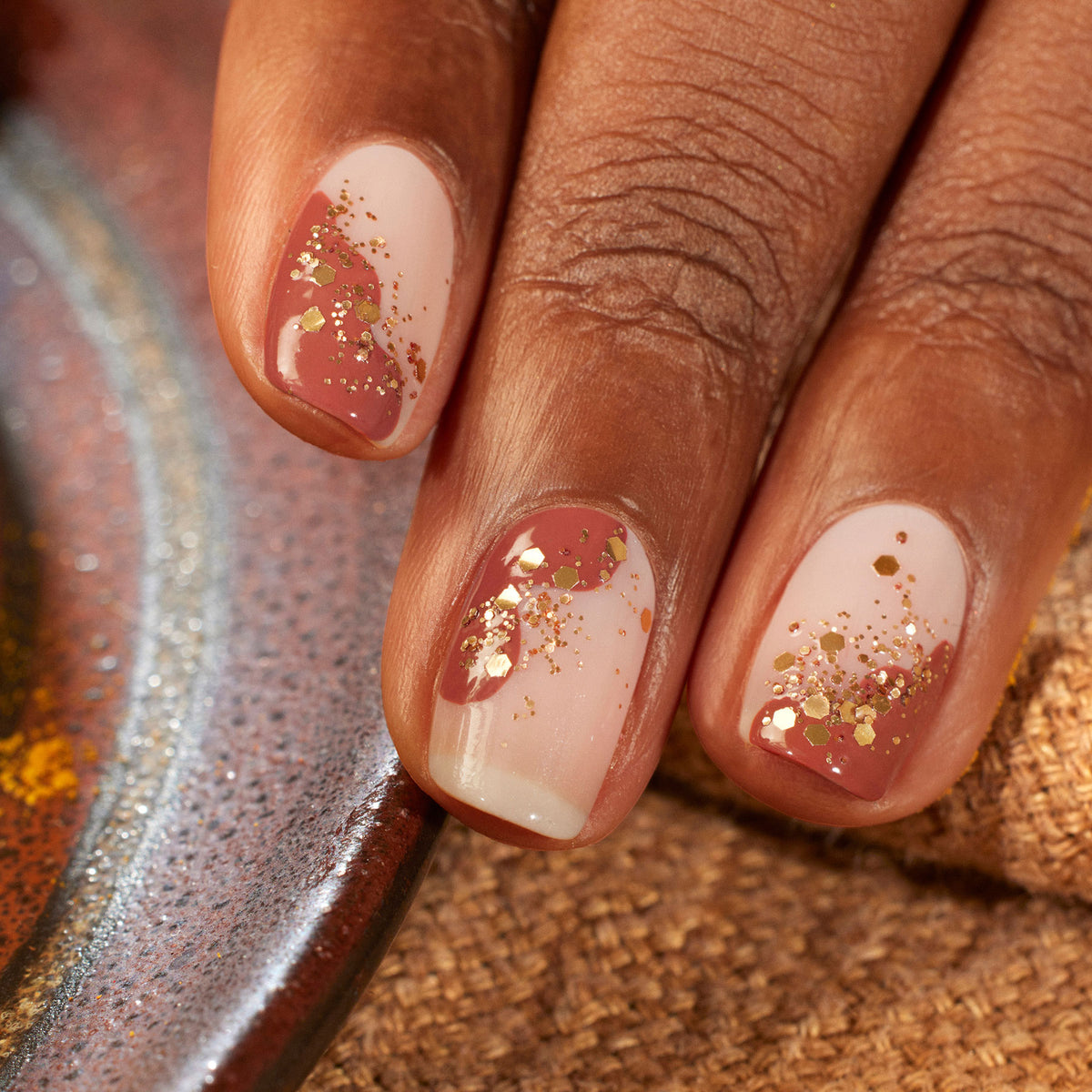 Une main à la peau brun moyen présente des ongles vernis rose nude, accentués de Paillettes Biodégradables Cuivrées de Manucurist près des pointes. L&