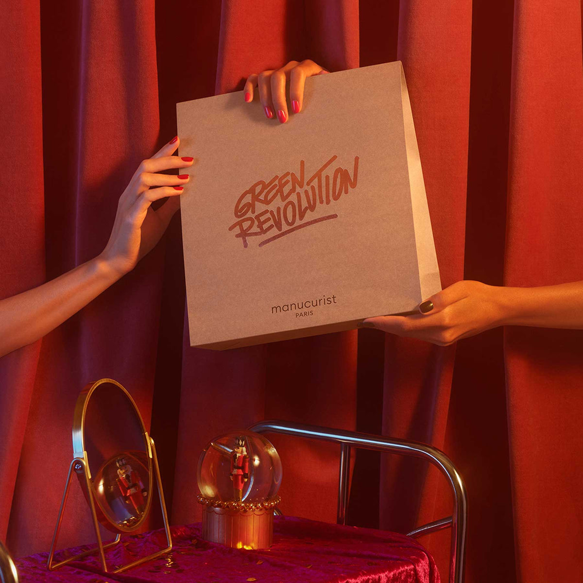 Two hands with red-painted nails hold a recyclable Medium Gift Bag by Manucurist. The background has red curtains, a small mirror, and a glass ornament placed on red velvet.