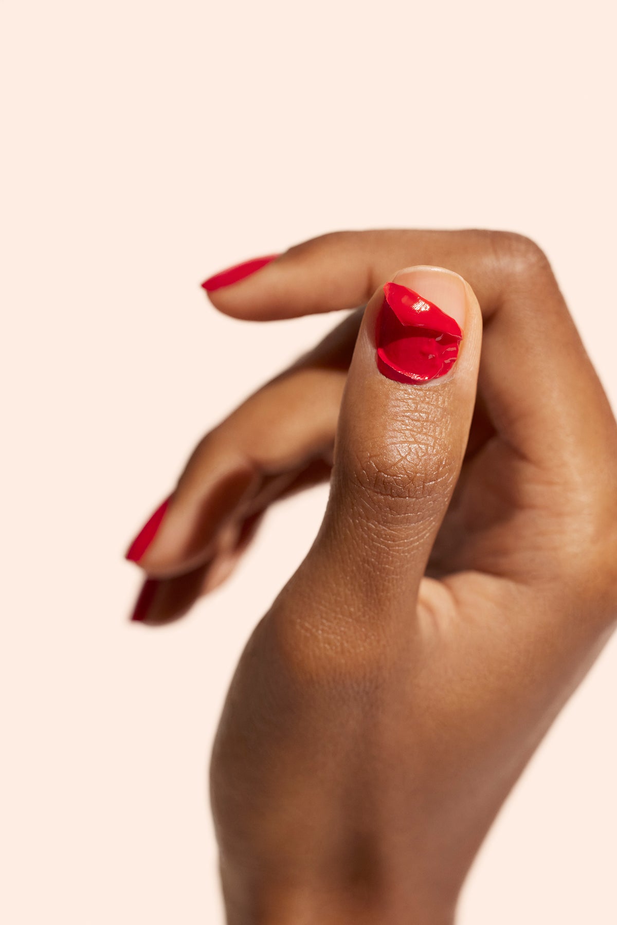 A hand with medium-brown skin holds up a thumb with chipped red nail polish. The other nails are also painted red. The background is a plain, light beige color.