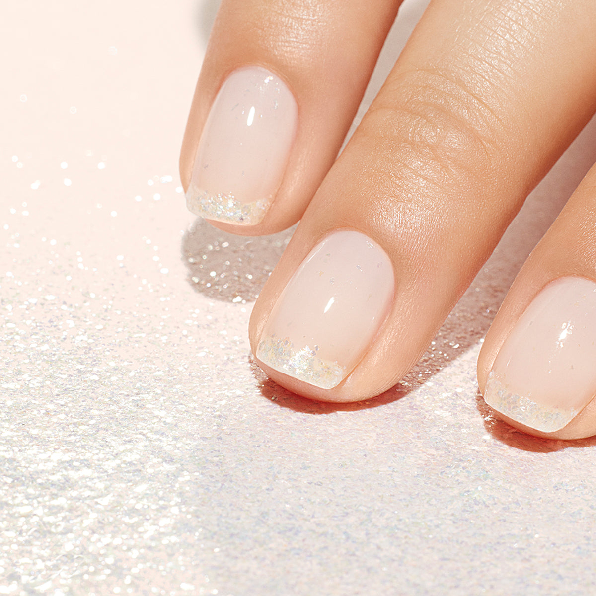Gros plan sur une main avec une manucure Manucurist Paillettes biodégradables Pearlescent ; ongles courts rose pâle ornés de délicates paillettes argentées et nacrées sur le bout des ongles. Un fond scintillant sublime ce look élégant.