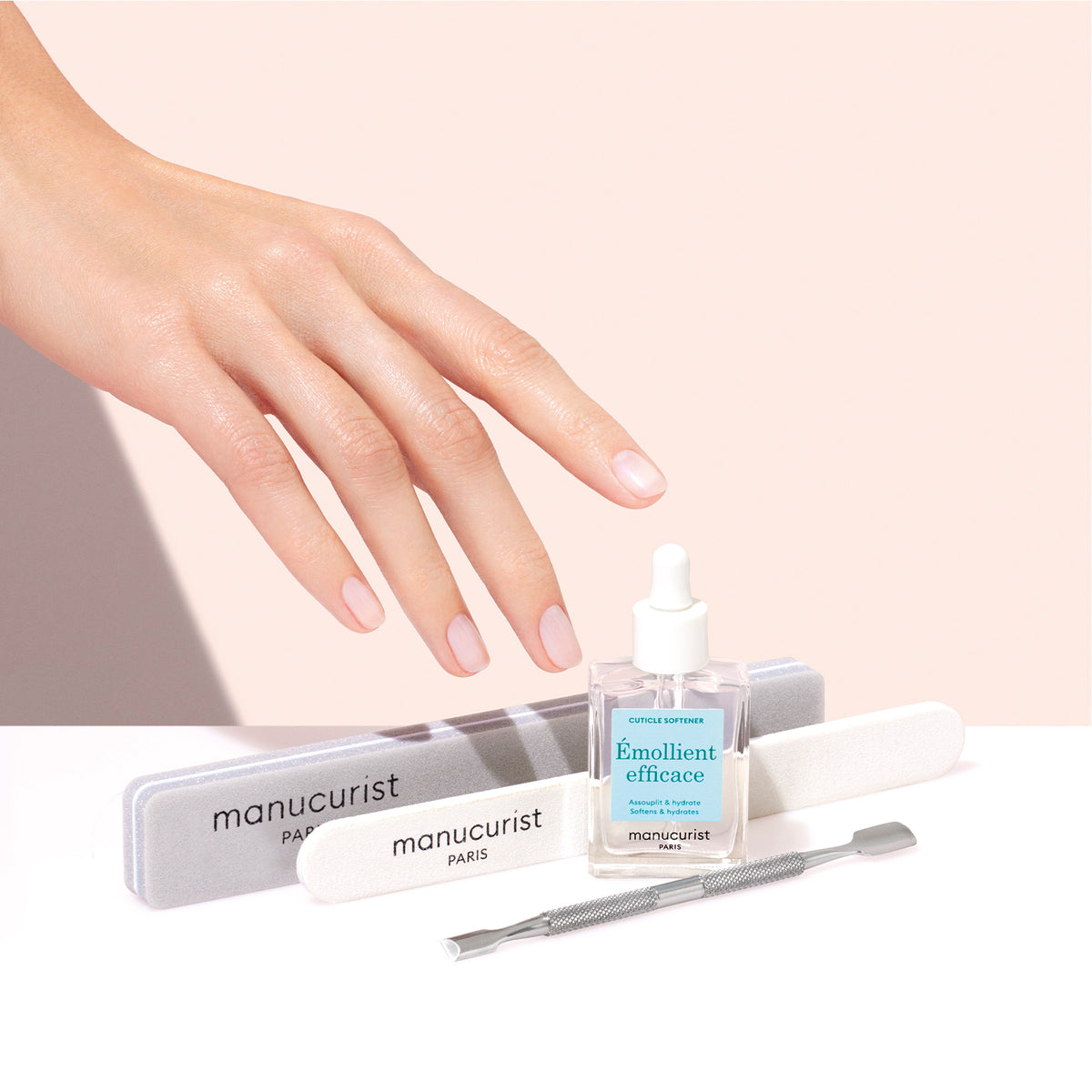 A hand with manicured nails hovers over Manucurist nail care essentials, including the Hydrating Cuticle Softener, emollient oil, nail files, and a cuticle pusher—all set against a soft pink background.