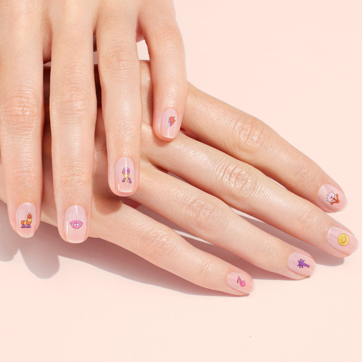 Gros plan sur des mains aux ongles courts et naturels, chacun décoré d’un Summer Nail Art Sticker ludique différent de Manucurist — smileys, lèvres et bien plus encore — sur une base rose clair pour une manucure amusante.