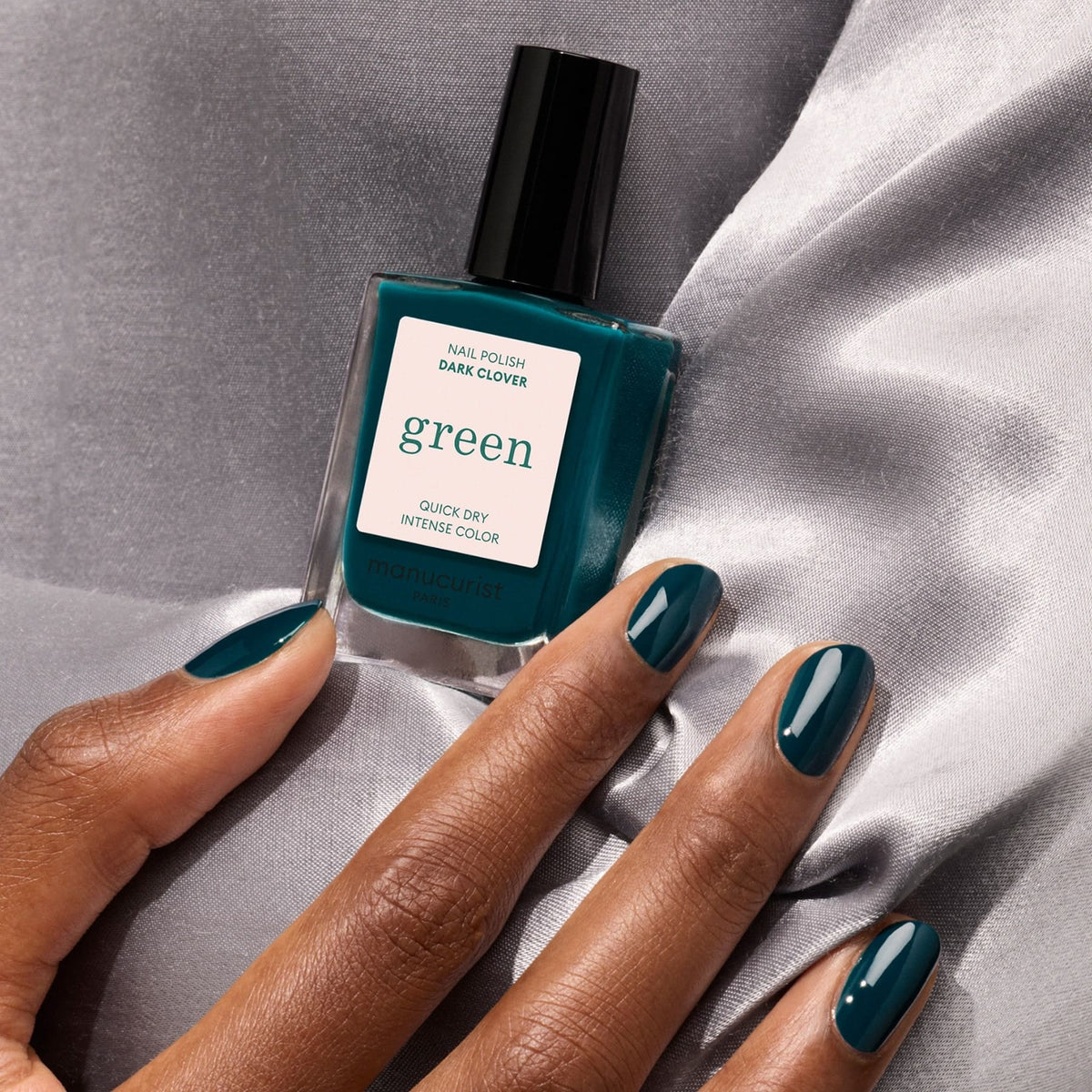 Une main aux ongles vert sarcelle foncé tient un flacon de vernis à ongles Manucurist Dark Clover, posée sur un tissu en satin gris.