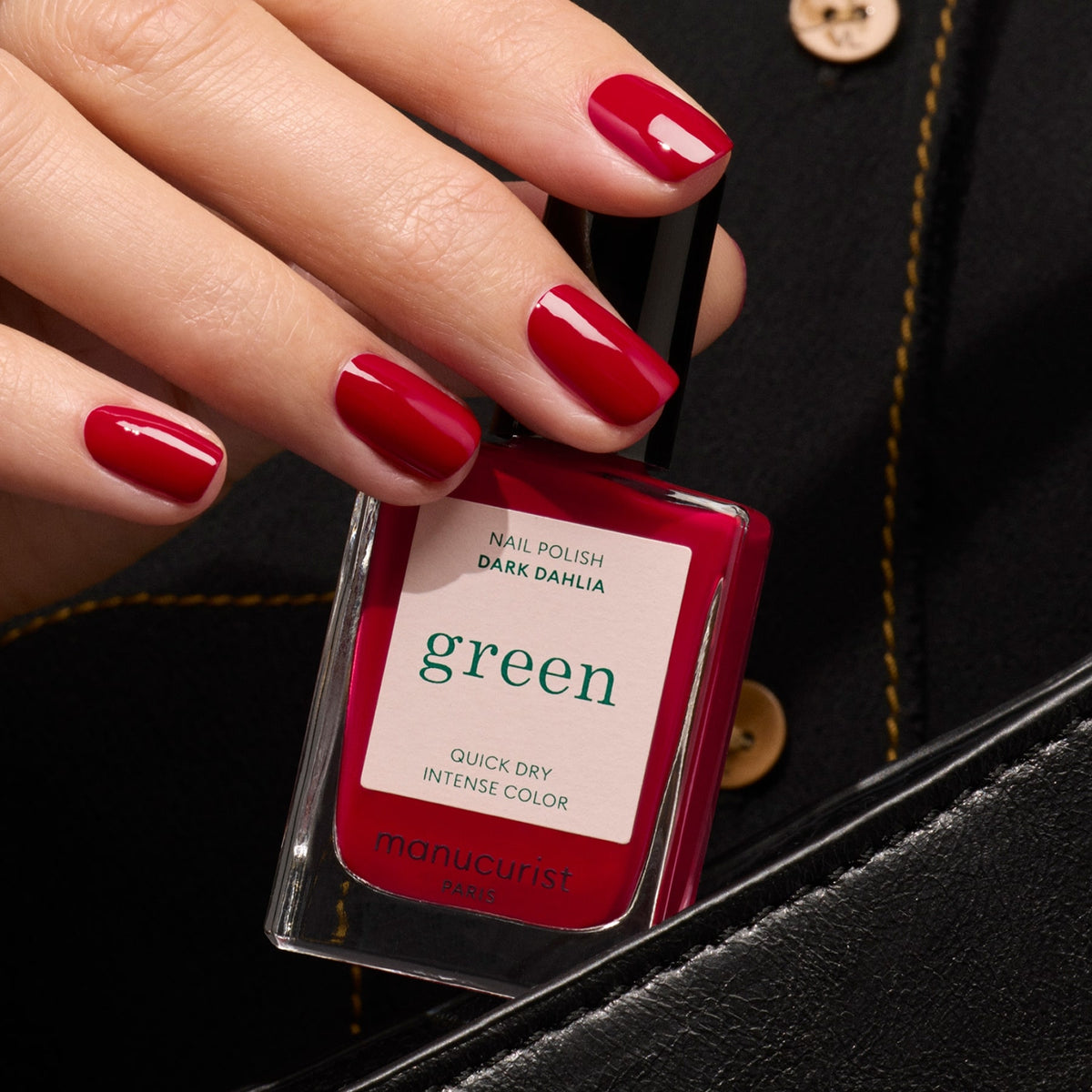 Une main aux ongles rouges brillants tient un vernis à ongles Manucurist Dark Dahlia — une teinte rouge foncé intense — devant une tenue noire à boutons dorés.