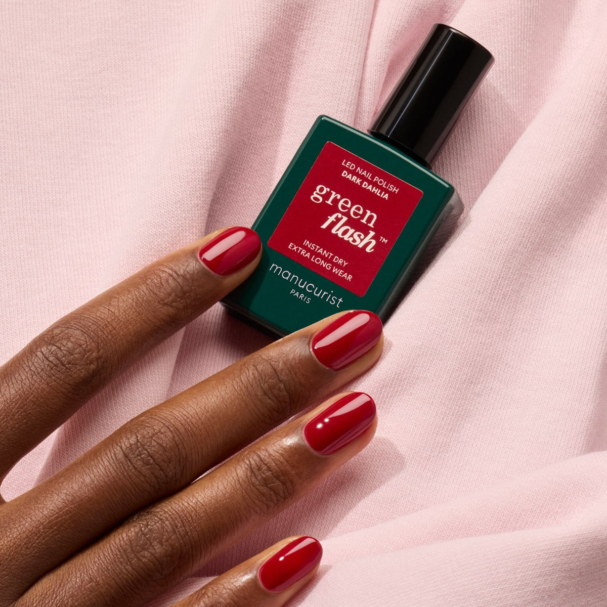 A hand with neatly manicured red nails holds a bottle of Manucurist Dark Dahlia gel polish against a soft pink fabric background.