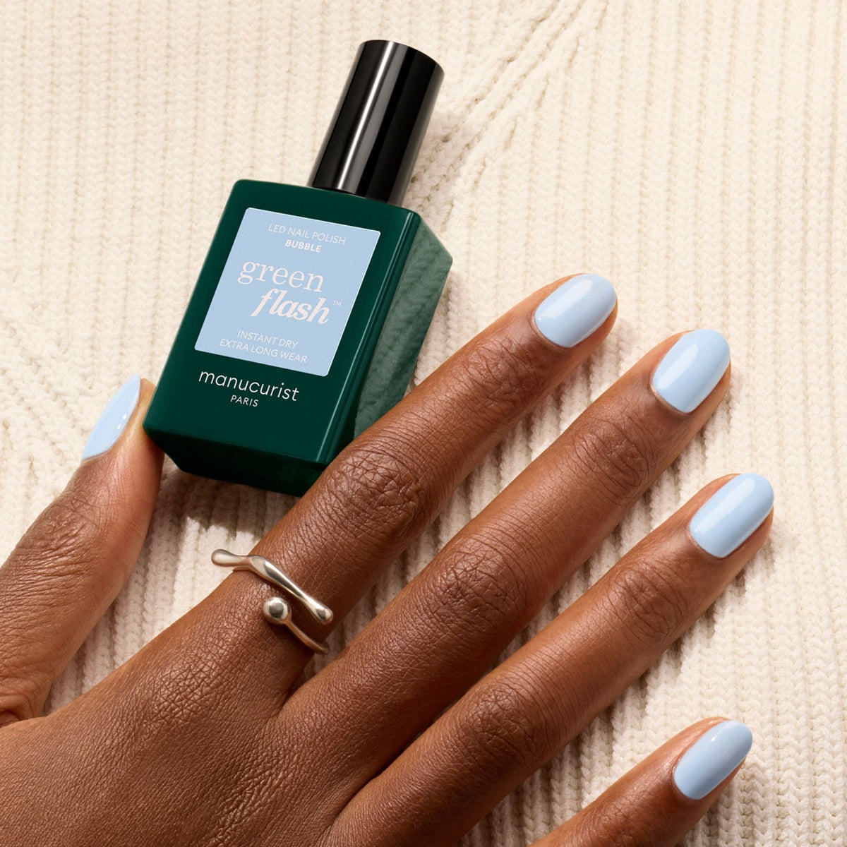 A hand with light blue Bubble LED Gel polish by Manucurist rests on cream knit fabric, adorned with a silver ring and holding a green bottle, highlighting subtle grey undertones.