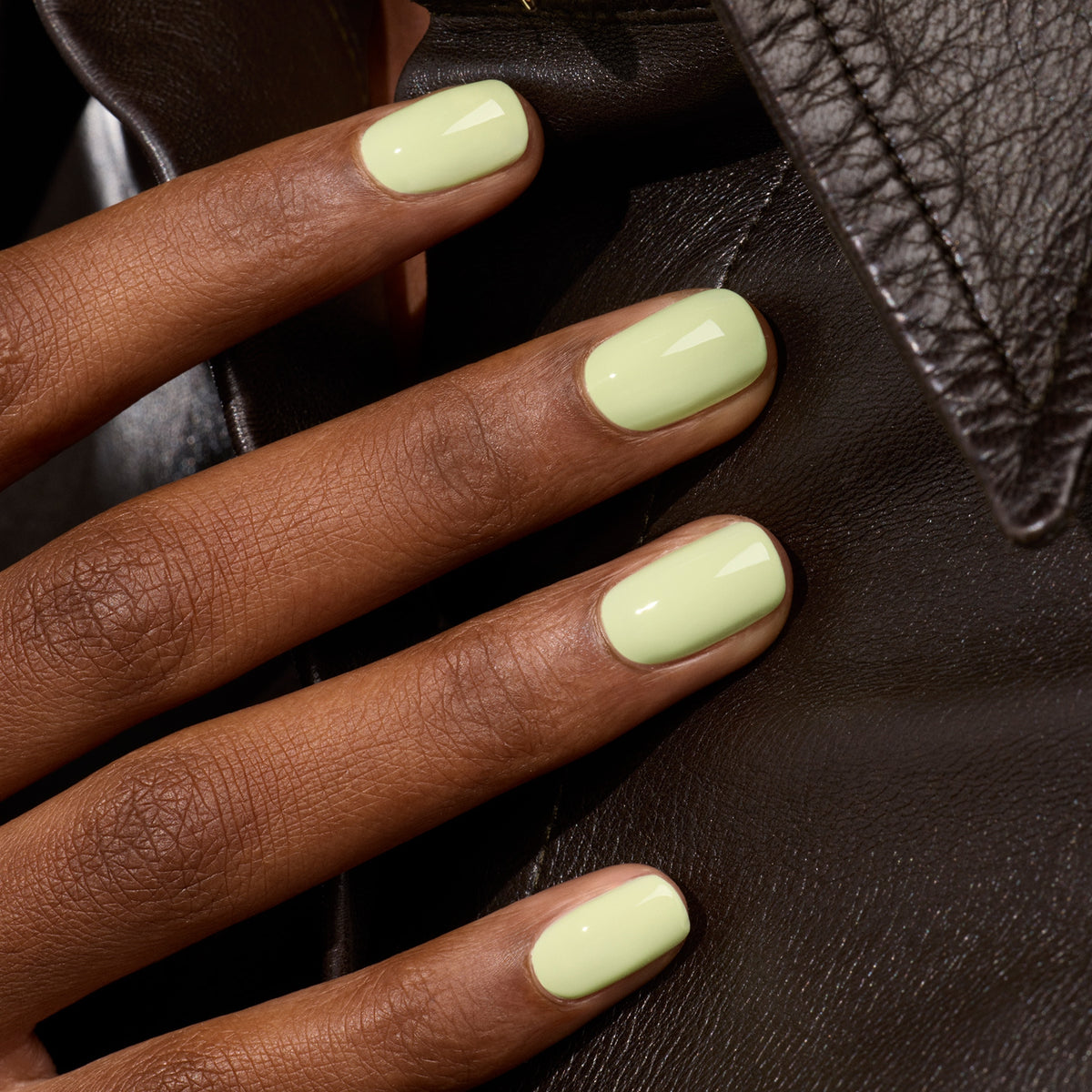 A hand with short, neatly filed nails painted in Pistachio, a glossy pastel yellow shade from Manucurist’s Green™ range, rests on a black leather jacket.