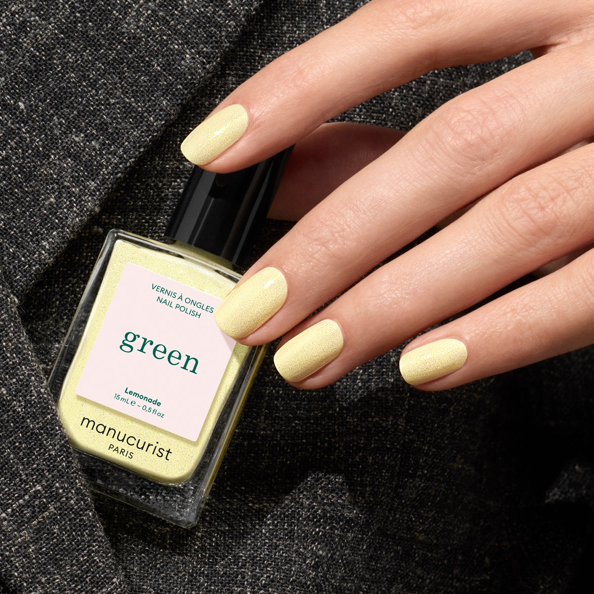 A hand with neatly manicured nails painted pale yellow holds a matching bottle of Lemonade by Manucurist, set against a textured dark fabric background.