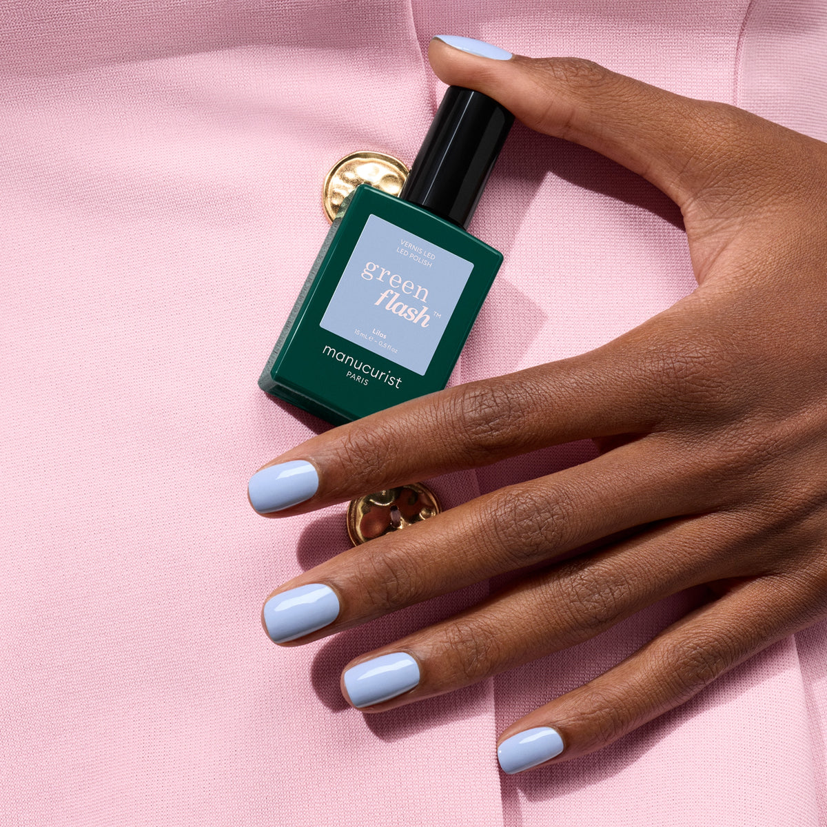 A hand with pastel blue gel polish holds a green bottle of Manucurist Lilas gel polish against a pale pink fabric background with a gold button.