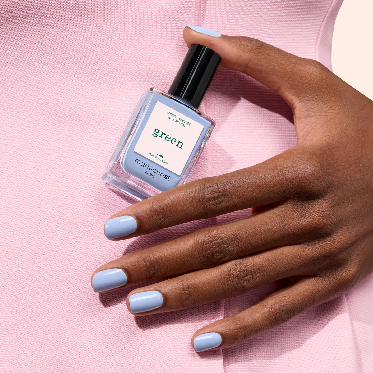 Une main avec du vernis à ongles bleu pastel tient un flacon de vernis à ongles Manucurist Lilas, sur un fond de tissu rose pâle.