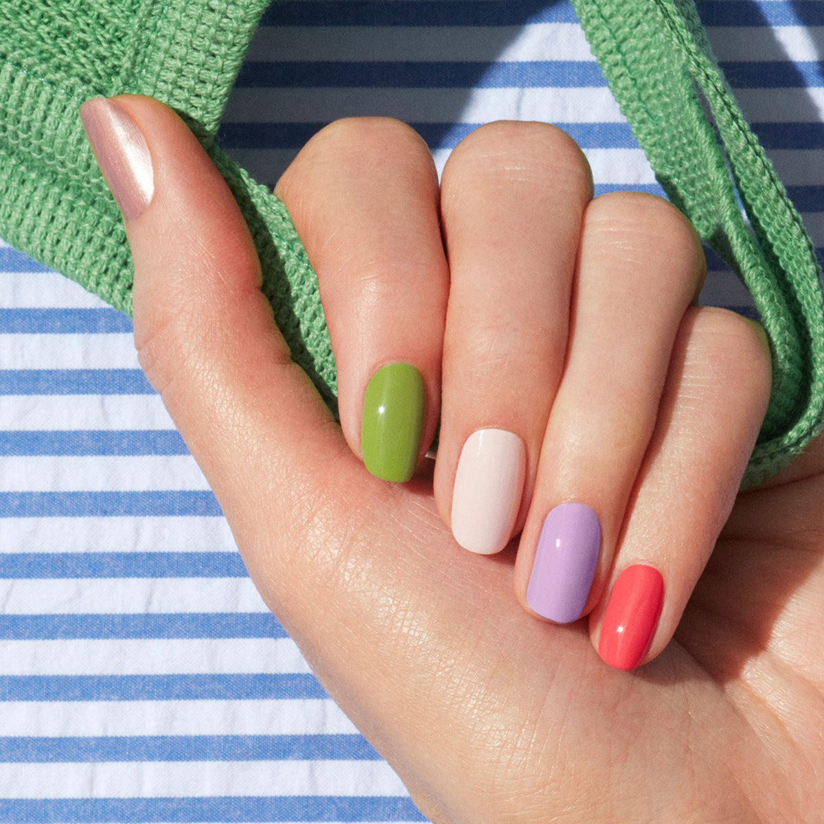 A hand with neatly manicured nails painted in five colors—metallic pink, green, white, lavender, and coral—rests on striped fabric with a green strap, highlighting playful nail art using Manucurist’s Dot it better!.