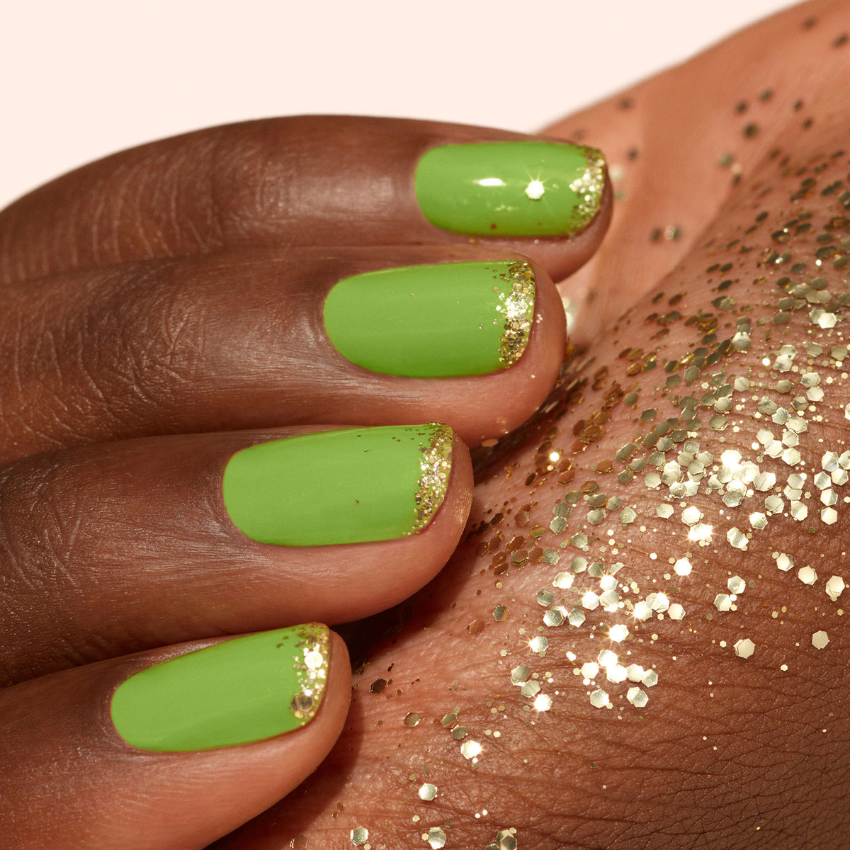 A hand with green gel polish and gold tips rests on skin sprinkled with Manucurist Gold Biodegradable Glitter, creating a neatly manicured look with festive, eco-friendly sparkle.