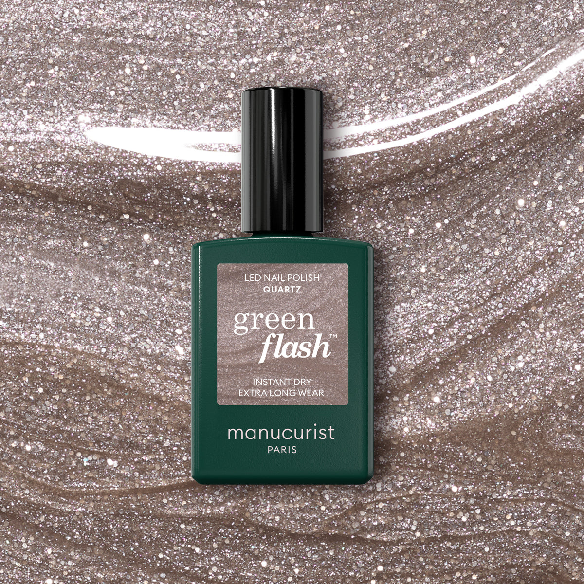 A bottle of Manucurist Quartz Green Flash semi-permanent nail polish sits on a glittery beige surface that matches the soft, refined pinkish-beige shade shown on the label.