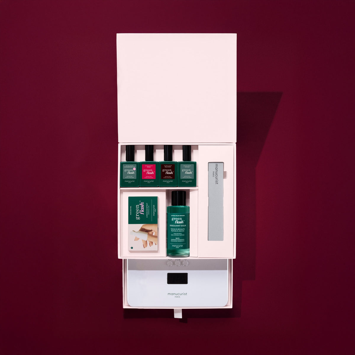 The Manucurist Starter Kit Gift Set (TEST COFFRET) comes in a pink box and includes green and red nail polish, a nail file, acetone-free remover pads, and a small gadget, all displayed against a dark maroon background.