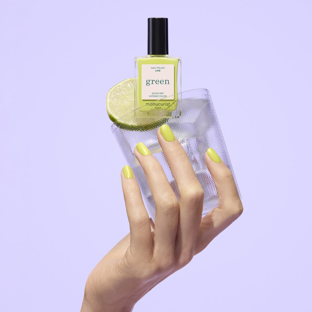 A hand with chartreuse green Manucurist Lime polish holds a glass of water with a lime slice, topped with a matching Manucurist Lime bottle, all set against a light purple background.