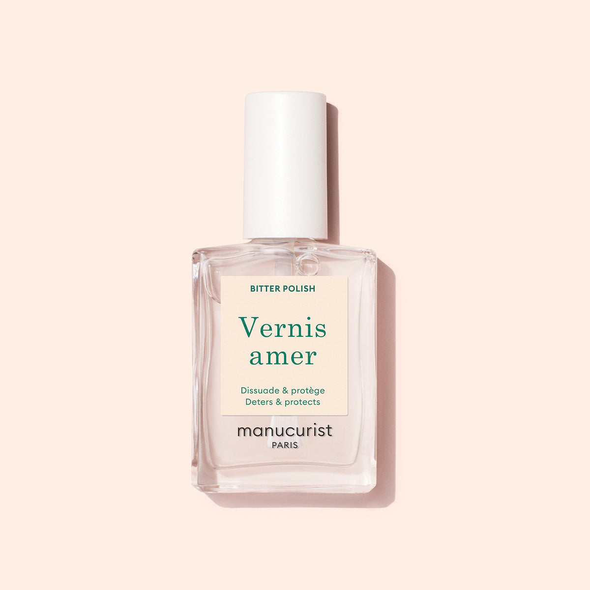 A clear rectangular bottle of Manucurist Bitter Polish with a white cap rests on a pale pink background. Its cream label with green and beige text highlights nail care benefits, helping prevent nail biting.