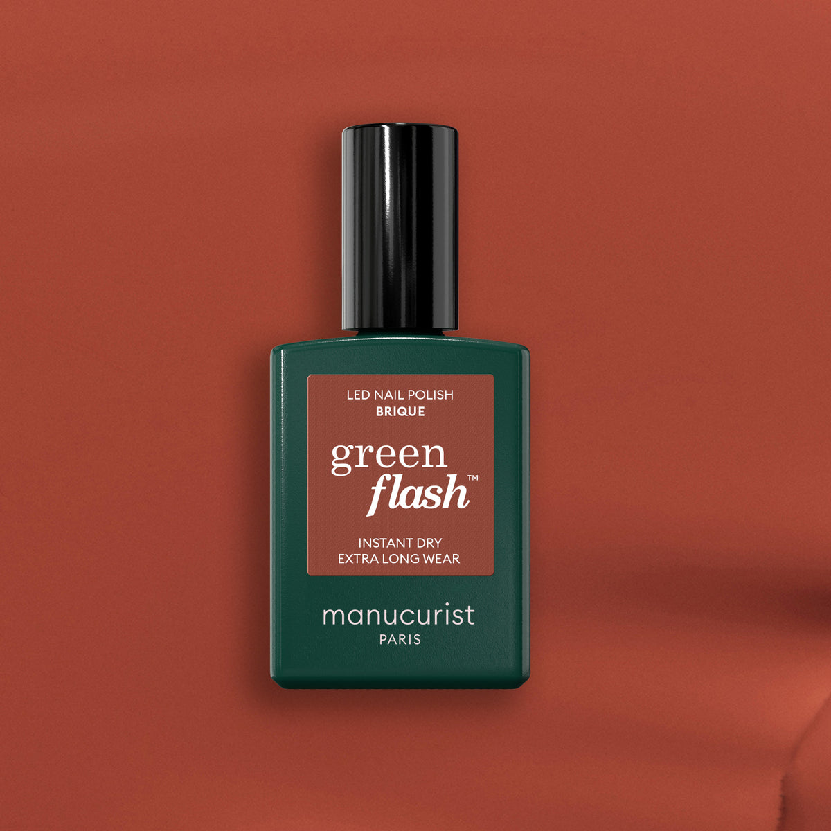 A bottle of Manucurist Brique Green Flash LED nail polish sits on a warm brown background. The dark green bottle with black cap and terracotta-accented label hints at the brick red shade inside.